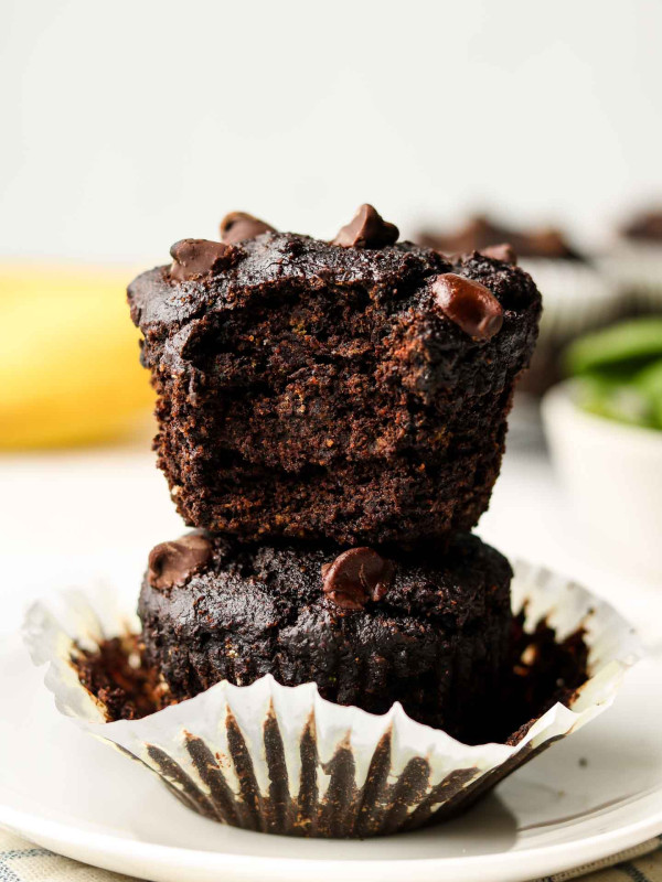 chocolate banana muffins stacked on each other with banana and spinach in background