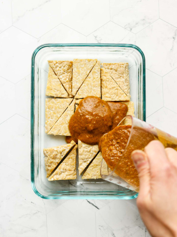 triangle tempeh in a meal prep bowl pouring marinade over