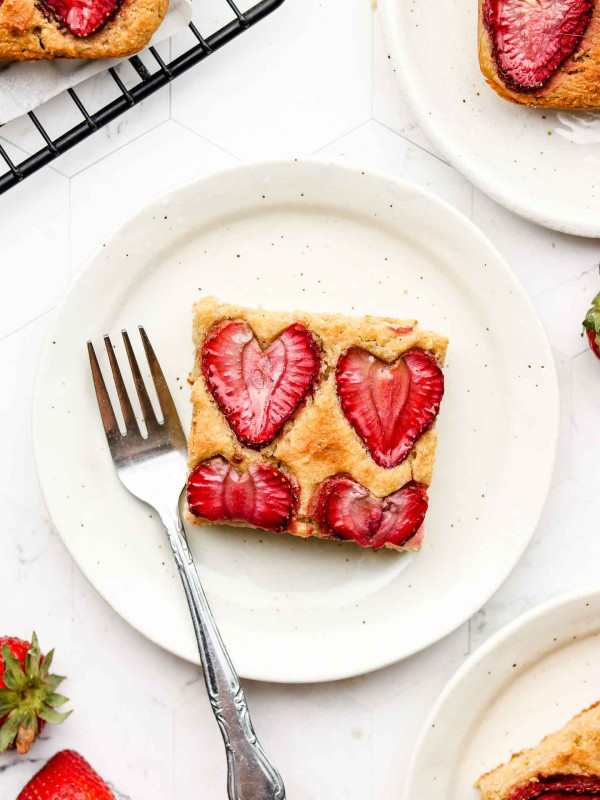 square piece of lemon sheet cake with strawberries on top on a white speckled plate and a fork on the side