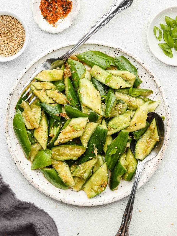 japanese smashed cucumber salad on a white speckled plate with toppings in white bowls and grey kitchen cloth in the corner