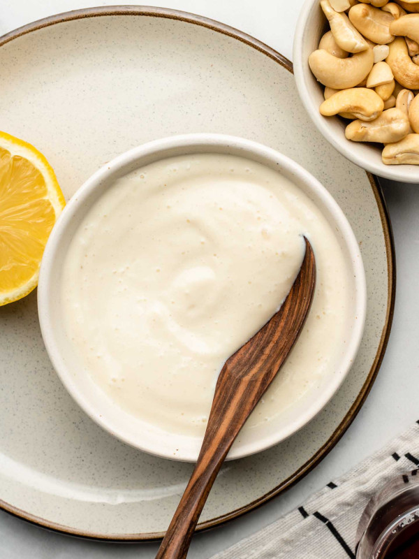 vanilla maple cashew cream in a white bowl with a wooden spoon inside and cashews on the top right corner