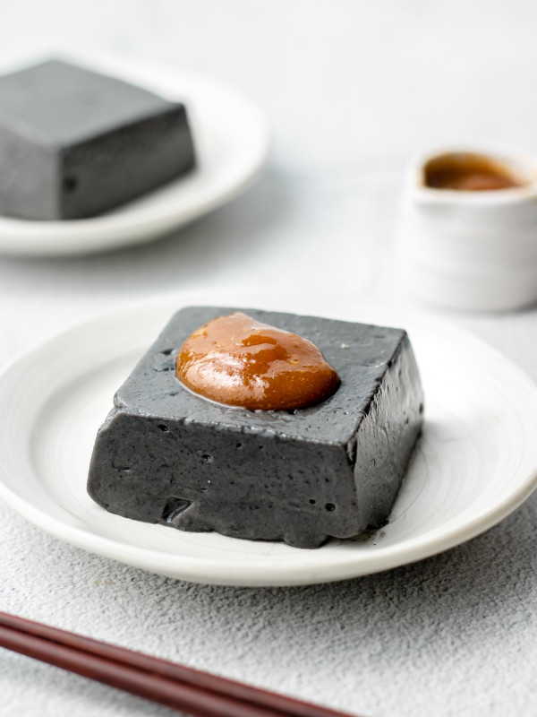 block of black sesame tofu on a white plate with miso sauce on top and sauce bottle behind it