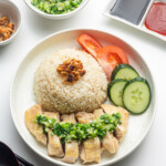 Shallow bowl of Hainanese chicken rice served with sliced chicken, cucumber slices, fluffy rice, and garnishes like scallion ginger sauce, light soy sauce, and chilli.