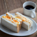 soft boileg egg sandwich on a white plate with black coffee beside it