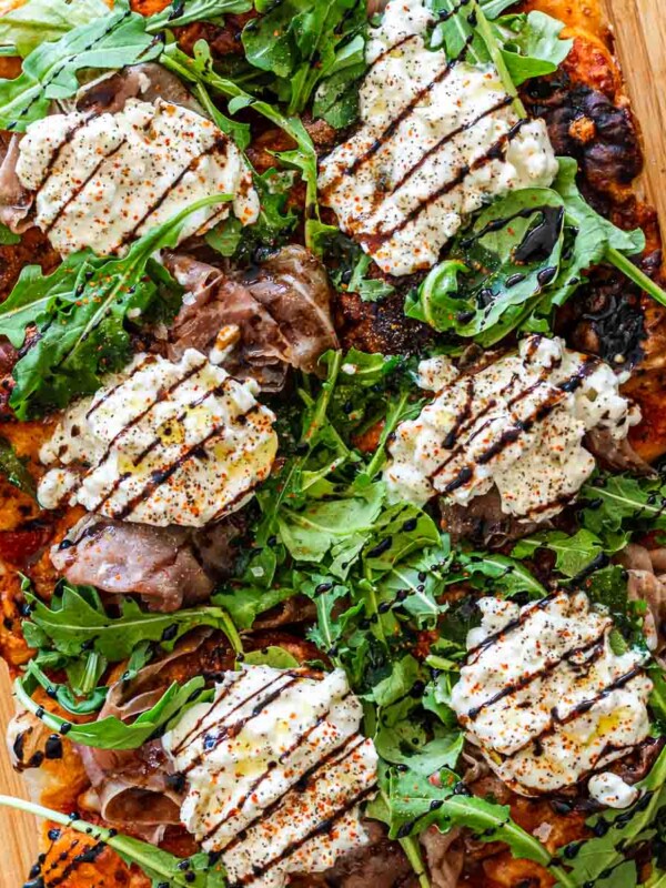 loaded focaccia pizza with prosciutto, burrata, arugula, tomatoes, mozzarella and balsamic glaze on a cutting board
