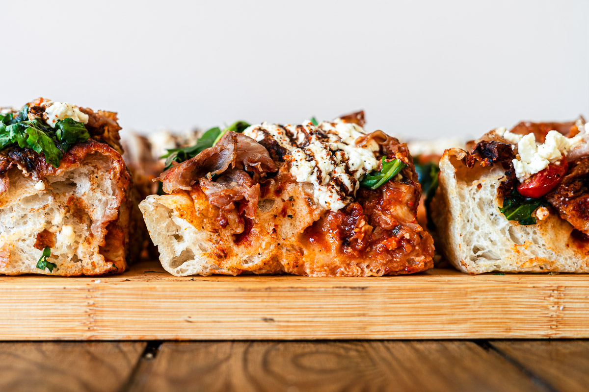 focaccia pizza slices topped with tomatoes, pizza sauce, arugula, burrata on a cutting board