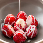 drizzling condensed milk over strawberries in a brown bowl