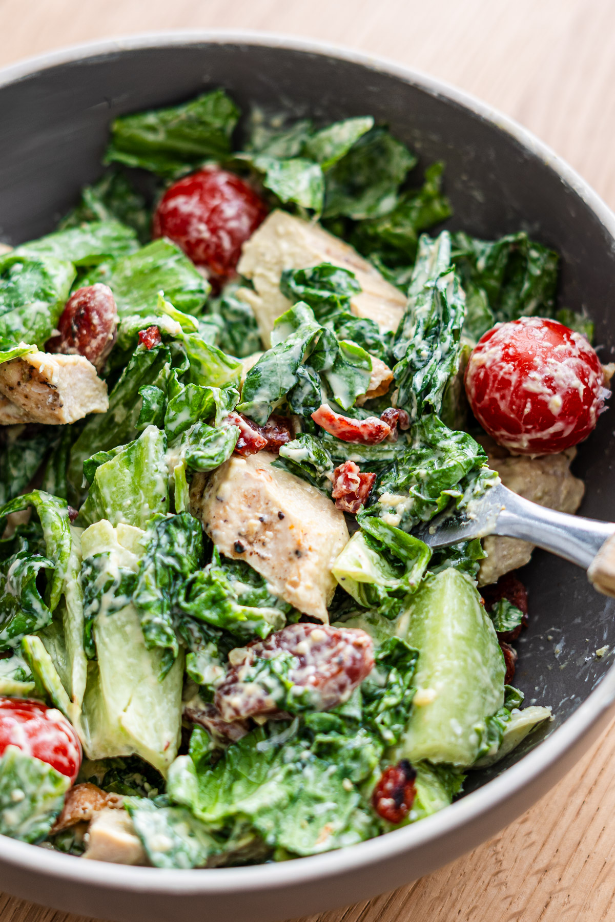 fork picking up caesar salad with lap cheong, lettuce, tomato, grilled chicken and fried wonton chilps in a brown bowl