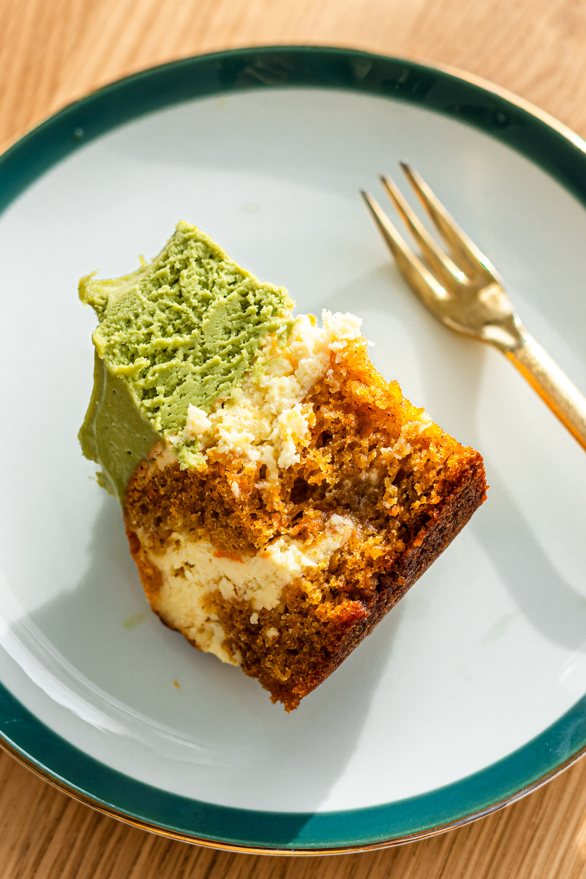 baked carrot cake layered with cheesecake topped with matcha cream cheese frosting on a green rimmed dessert plate with a bite taken out of it