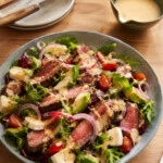 steak salad with medium rare steak slices, brie, spring mix, avocado, tomato, red onion topped with creamy japanese sesame dressing in a ceramic grey bowl with a side of dressing in a green bowl on the side and side dishes