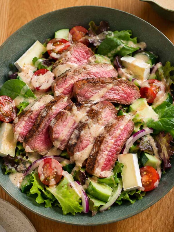 steak salad with slices of rare steak, brie, spring mix, cucumber, tomato, red bell pepper in a blue-grey tone shallow bowl