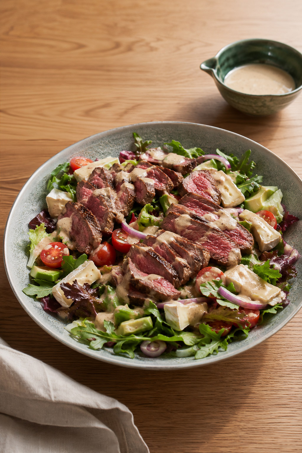 rare steak steak salad with spring mix, brie,avocado, tomato, red onion topped with creamy japanese sesame dressing in a ceramic grey bowl with a side of dressing in a green bowl on the side